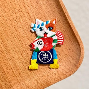 May include: Embroidered patch featuring a white, orange, and blue cat holding a red and white fish. The cat wears a blue and yellow outfit with Japanese text. The patch is on a wooden surface.