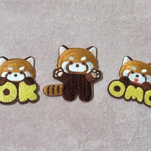 May include: Three embroidered patches featuring red pandas. One red panda holds the word "OK" in yellow, another stands with arms outstretched, and the third holds "OMG" in yellow. The patches have brown, white, and yellow details.