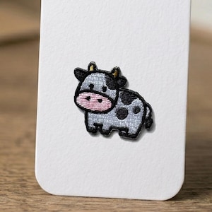 May include: A small embroidered patch featuring a cartoon cow with light grey fur, black spots, and a pink snout. The cow has small horns and is outlined in black thread, presented on a plain white rectangular card backing.