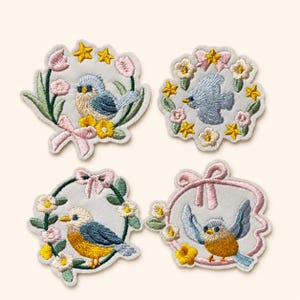 May include: Four embroidered fabric patches featuring birds surrounded by floral wreaths, bows, and stars. The designs include blue and yellow birds, pink and white flowers, green leaves, and yellow stars on a light gray background fabric.