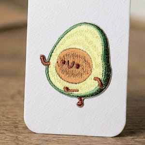 Puede incluir: Parche bordado de aguacate con una cara sonriente, con cáscara verde, pulpa amarilla y un hueso marrón. El aguacate tiene brazos y piernas pequeños, lo que le da una apariencia juguetona. El parche está sobre un fondo blanco.