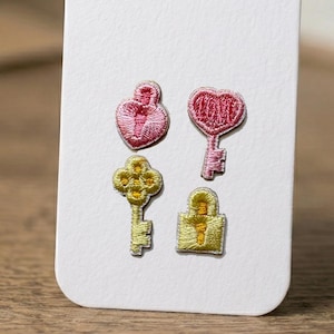 May include: Four embroidered patches: a pink heart-shaped lock, a pink heart-shaped key, a yellow key, and a yellow lock. The patches are on a white background. The patches are likely for clothing or craft projects.