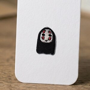 May include: A small embroidered patch featuring a character with a white mask, black body, and red markings around the eyes and mouth. The patch is attached to a white rectangular card with rounded corners, displaying detailed thread work.