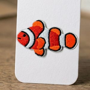 May include: Embroidered clownfish patch. The patch has a white body with orange and black detailing. The fish is shown in profile, with its fins and tail clearly visible. The patch is designed for application on clothing or accessories.