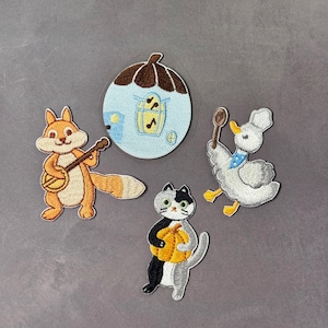 May include: Embroidered patches featuring a squirrel playing a banjo, a blue house with musical notes, a duck holding a spoon, and a cat holding a pumpkin. The patches have detailed stitching and vibrant colors.