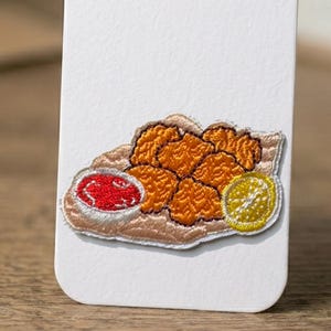 May include: Embroidered patch featuring a plate of fried chicken nuggets, a small bowl of red dipping sauce, and a lemon slice. The patch has a brown, orange, red, and yellow color scheme. The patch is on a white background.