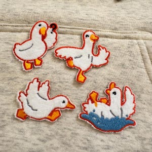 May include: Four embroidered patches of white geese with orange beaks and feet, outlined in red. One goose is swimming in blue water. The patches are on a light grey fabric background.