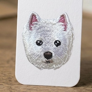 May include: Embroidered patch featuring a West Highland White Terrier dog. The dog's face is white with black eyes and nose, and pink accents in the ears. The patch is on a white background.