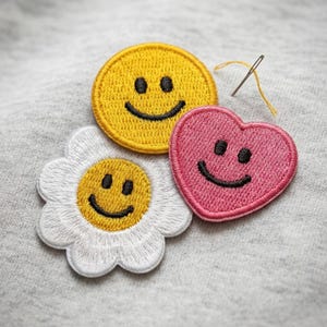 May include: Embroidered patches in the shape of a yellow smiley face, a pink heart, and a white daisy with a yellow center. Each patch has a black smiling face. A needle with yellow thread is also visible.