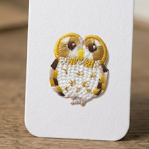 May include: Embroidered owl patch in gold, brown, and white. The owl is on a white background. The owl's design features detailed stitching, creating a textured appearance. The patch is likely for clothing or accessories.