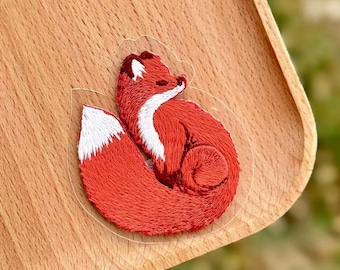 Op de afbeelding: Geborduurde patch met een opgerolde vos. De vos is voornamelijk rood met een witte borst en staartpunt. De patch zit op een doorzichtige achtergrond, geplaatst op een houten oppervlak.
