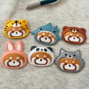 Peut inclure: Écussons brodés représentant un panda roux déguisé en différents animaux : tigre, requin, ours, lapin, panda et loup. Chaque écusson a un visage blanc et est bordé d'une couleur assortie. Les écussons sont sur un fond de tissu gris.