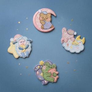 May include: Four embroidered patches with a bedtime theme are arranged on a blue background. The patches feature a bear on a crescent moon, a sheep, a rabbit on a cloud, and a frog, all in pastel colors. Small gold stars are scattered around the patches.