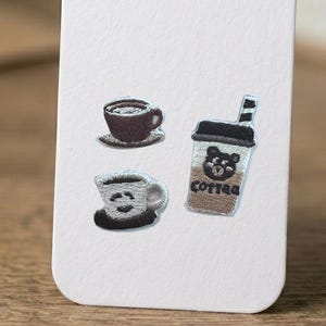 May include: Three embroidered patches featuring coffee-themed designs: a dark brown coffee cup on a saucer, a light-colored cup with a dark base, and a tall coffee cup with a straw, a bear face graphic, and the text "coffee" in black thread.