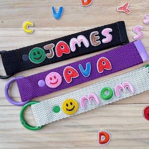 May include: Three colorful keychains with embroidered names "JAMES", "AVA", and "MOM" with a smiley face patch on each. The keychains are made of black, purple, and white canvas.