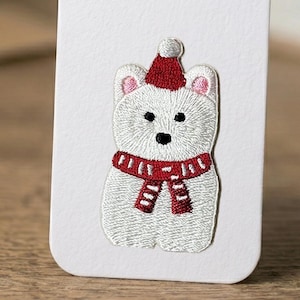 May include: Embroidered patch of a white polar bear wearing a red Santa hat and scarf. The bear has black eyes and nose, and pink inner ears. The scarf has a repeating pattern. The patch is on a white background.