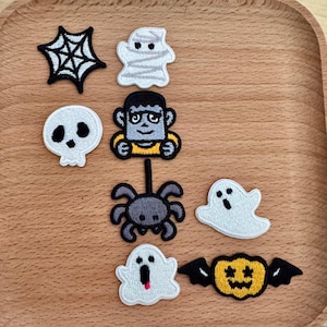 May include: A collection of Halloween-themed embroidered patches. Designs include a spider web, mummy, skull, Frankenstein's monster, spider, ghost, and jack-o'-lantern with bat wings. The patches are in various colors, including black, white, gray, and yellow.