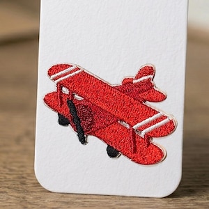 Puede incluir: Un parche bordado de avión biplano rojo con rayas blancas y detalles negros sobre un fondo blanco. El avión tiene una hélice y un diseño detallado. Adecuado para ropa o accesorios.