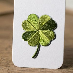 Puede incluir: Un trébol de cuatro hojas bordado en tonos verdes, fijado a una tarjeta blanca. Las hojas del trébol tienen costuras intrincadas y el tallo es de un verde más oscuro. La tarjeta tiene esquinas redondeadas.