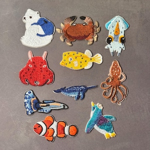 May include: A collection of embroidered patches featuring various sea creatures. Includes a polar bear with a penguin, a crab, a squid, an octopus, a clownfish, a yellow fish, a blue whale, a seahorse, and a penguin.