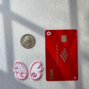 May include: A red Bank of America Visa credit card, a quarter, and two embroidered white and pink butterfly wing patches are arranged on a white surface. The card has the Visa logo and the Bank of America logo.
