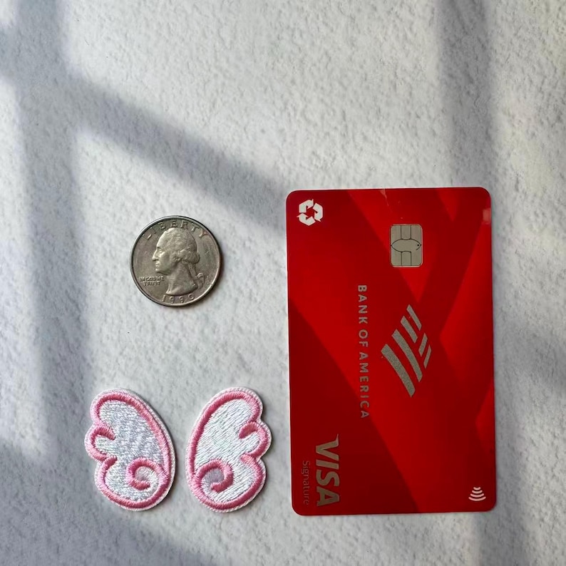 May include: A red Bank of America Visa credit card, a quarter, and two embroidered white and pink butterfly wing patches are arranged on a white surface. The card has the Visa logo and the Bank of America logo.