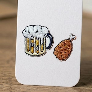 以下が含まれることがあります： 白い泡のビールジョッキとフライドチキンの脚が描かれた刺繍パッチ。 ビールジョッキは黄色で、鶏の脚は茶色です。 衣類やアクセサリーに加えるのに最適です。