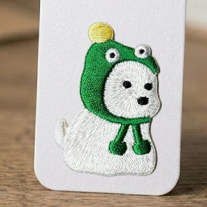May include: Embroidered patch featuring a white dog wearing a green frog hat with a yellow pom-pom. The patch has black eyes and nose, and green accents on the hat and scarf. The patch is on a white background.