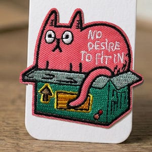 Puede incluir: Parche bordado con un gato rosa con ojos ribeteados en blanco y el texto "NO DESIRE TO FIT IN". El gato está en una caja verde con una flecha amarilla. El parche tiene un borde negro y está sobre un fondo blanco.