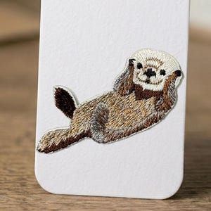 May include: Embroidered patch featuring a whimsical otter design. The otter is depicted in shades of brown, tan, and black, with detailed stitching. The patch is likely for clothing or accessories, adding a touch of nature-inspired charm.