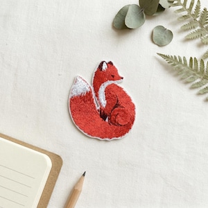 Pode incluir: Remendo bordado de uma raposa enrolada. A raposa é principalmente vermelha com a cauda com ponta branca e detalhes brancos. O remendo está em uma superfície creme, com um caderno, um lápis e galhos de vegetação no fundo.