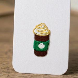 Puede incluir: Un parche bordado que muestra una taza de café marrón con una funda verde y un área de logotipo circular blanca. La taza está cubierta con un remolino de crema batida amarilla. El diseño está cosido sobre un soporte de tarjeta blanca.