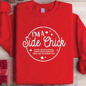 以下が含まれることがあります： 白い文字で「I'M A Side Chick」と書かれた赤いスウェットシャツ。テキストの下には、おかずのリストがあります。スウェットシャツは木製の表面に折りたたまれており、松ぼっくりやオーナメントなどの休日の装飾が施されています。
