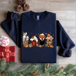May include: Navy blue sweatshirt with a graphic of four mugs with autumnal designs. The mugs are decorated with characters from the Harry Potter series, including Harry Potter, Hermione Granger, Ron Weasley, and a Hogwarts house crest. The text "G" is visible on one of the mugs.