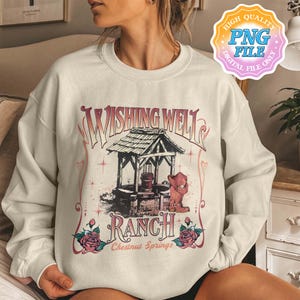 Puede incluir: Sudadera crema con un gráfico de estilo vintage de un pozo de los deseos, el texto "Wishing Well Ranch Chestnut Springs", rosas y una bota de vaquero. El diseño es en tonos rojos y marrones.