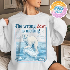 Pode incluir: Camisola branca com um gráfico de um urso polar num icebergue a derreter, com o texto "The wrong ice is melting" em azul e vermelho. O design inclui água azul, nuvens e estrelas.