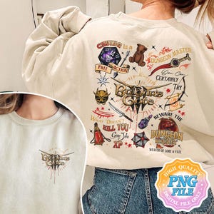 May include: A beige sweatshirt with a graphic print of a Dungeons and Dragons themed design. The design includes a dice, a teddy bear, a sword, and the text "Crying is a Dungeon Master You Can Certainly Try Baldur's Gate What Doesn't Kill You Give You XP Beware the Dungeon Master Weaver of Lore & Fate".
