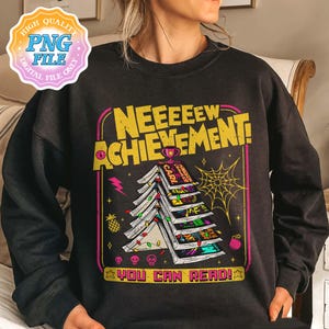 May include: Black sweatshirt with a graphic that reads "NEEEEEW ACHIEVEMENT!" in yellow and pink. The design features a stack of books shaped like a Christmas tree, with the text "YOU CAN READ!" at the bottom.