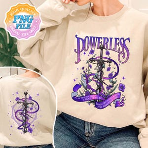 May include: A cream-colored sweatshirt with a purple and black graphic design featuring a sword, flowers, and the text "POWERLESS". The text below the graphic reads "IF I AM TO BE YOUR ENEMY, I WANT IT TO BE BECAUSE SHE LOATHES HERSELF FOR WANTING ME."