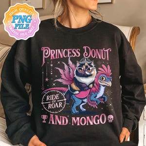 May include: A black sweatshirt with a whimsical graphic featuring a cat wearing glasses and a crown, riding a blue and pink dinosaur-like creature. The text reads "Princess Donut" and "Ride or Roar and Mongo."