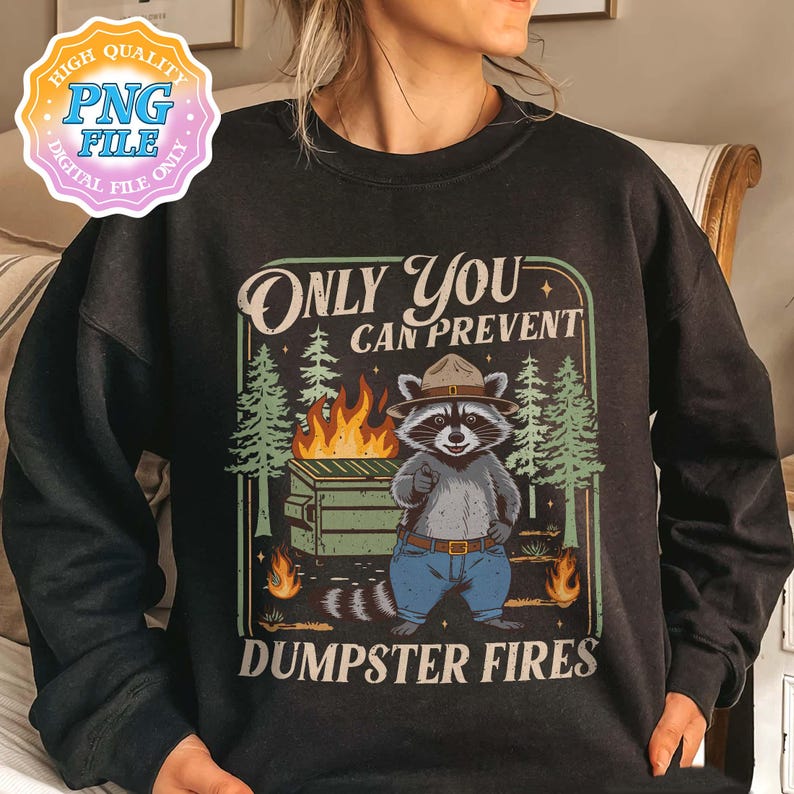May include: Dark gray sweatshirt with a graphic of a raccoon in a ranger hat, pointing at a dumpster fire. The text reads "Only You Can Prevent Dumpster Fires." The design includes trees and flames.