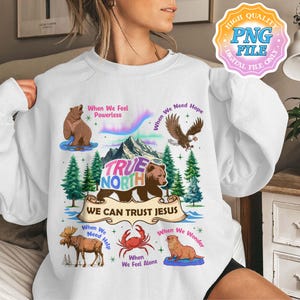 May include: White sweatshirt with a vibrant graphic of various animals, including a bear and eagle, with the text "TRUE NORTH" and "WE CAN TRUST JESUS". The design is set against a backdrop of mountains and trees. The sweatshirt has a crew neck.