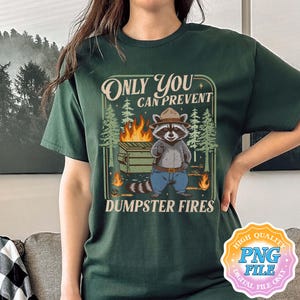 May include: Dark green t-shirt featuring a raccoon in a ranger hat, standing in front of a dumpster fire. The text reads "Only You Can Prevent Dumpster Fires". The design includes trees and flames.