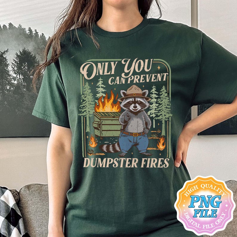 May include: Dark green t-shirt featuring a raccoon in a ranger hat, standing in front of a dumpster fire. The text reads "Only You Can Prevent Dumpster Fires". The design includes trees and flames.
