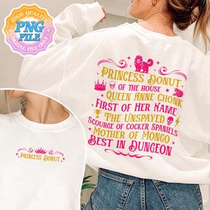 May include: White sweatshirt with a pink and gold graphic. The graphic reads "PRINCESS DONUT" and other phrases. A crown, skull, and dog are also featured in the design. The sweatshirt is a PNG file.