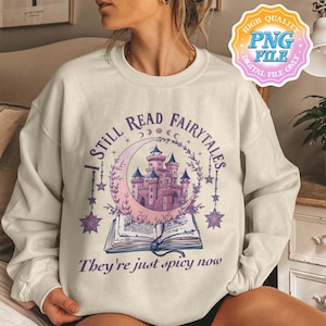 Puede incluir: Una sudadera de color beige claro con un diseño gráfico de una luna creciente, un castillo y un libro abierto. El texto "I still read fairytales they're just spicy now" está impreso en la sudadera.