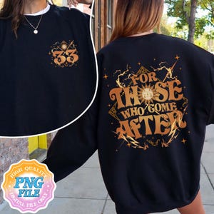 Puede incluir: Sudadera negra con texto y gráficos dorados. La parte delantera presenta el número "33" con diseños de sol y estrellas. La parte trasera muestra la frase "FOR THOSE WHO COME AFTER" en una fuente estilizada con acentos celestiales.