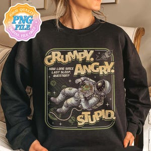 May include: Black sweatshirt with a retro-style graphic. The design features an astronaut in a spacesuit with the words "GRUMPY, ANGRY, STUPID" and the question "HOW LONG SINCE LAST SLEEP, QUESTION?" in a green and yellow color scheme.