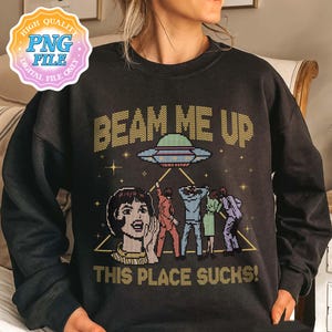 May include: Dark gray sweatshirt with the text "BEAM ME UP THIS PLACE SUCKS!" and a pixelated graphic of a UFO and people being beamed up. The design features retro-style illustrations in shades of yellow, red, blue, and green.