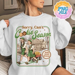 Könnte beinhalten: Weißes Sweatshirt mit einem Aufdruck „Sorry. Can't. Cookie Season“. Das Design zeigt zwei Mädchen in grünen Uniformen, die mit einem Wagen voller Kekse gehen. Ein Keks und ein Haus sind ebenfalls im Design enthalten.